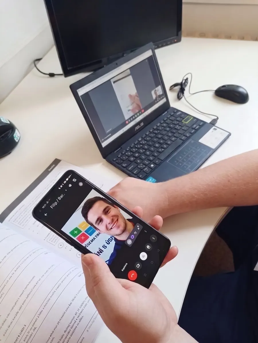Online hodina s lektorem Doučse: výklad běží v mobilu, vedle je notebook a pracovní sešit.